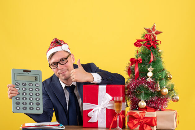 专业人士帅哥在圣诞树旁摆着竖起大拇指的牌子 黄色的礼物摆在桌子上办公室坐着礼物