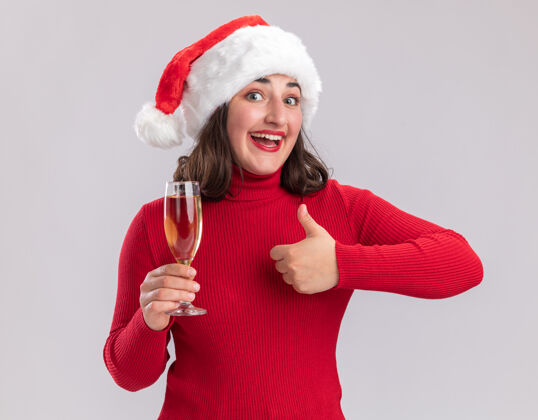 抱着穿着红色毛衣 戴着圣诞帽的快乐小女孩 手里拿着一杯香槟看着镜头 兴高采烈地微笑着 竖起大拇指站在白色背景上高兴微笑玻璃杯
