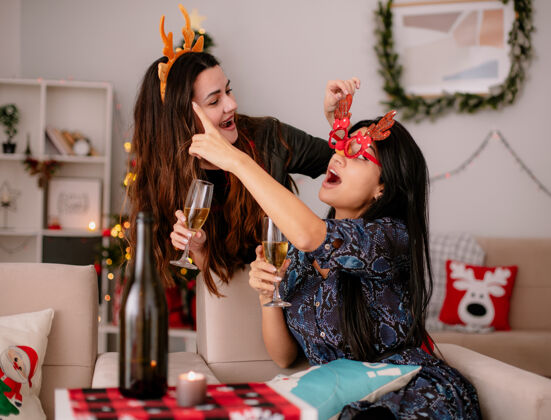 年轻戴着驯鹿头带的快乐的年轻女孩拿着一杯香槟 看着戴着驯鹿眼镜的朋友指着坐在扶手椅上的一边 在家里享受圣诞节的时光香槟看朋友