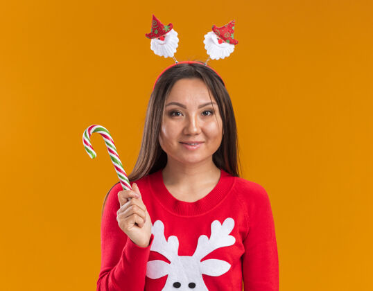 毛衣高兴的年轻亚洲女孩戴着圣诞发箍和毛衣拿着圣诞糖果隔离在橙色背景亚洲人头发年轻