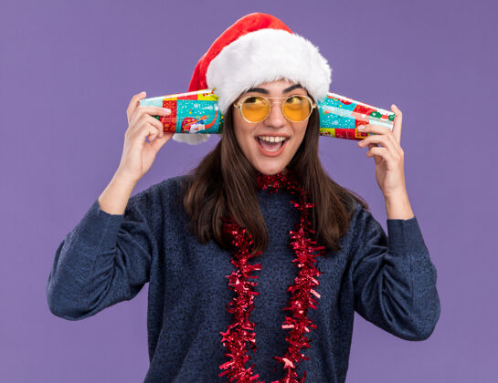 耳朵戴着太阳眼镜 戴着圣诞帽 脖子上戴着花环的快乐的年轻白种人女孩拿着纸杯 紧挨着耳朵 在紫色的背景上隔离开来 留着复印空间关闭眼镜帽子