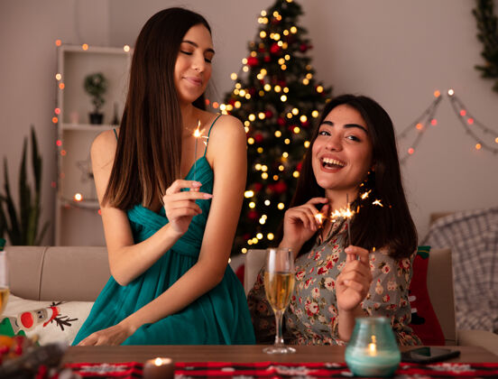 高兴高兴的年轻女孩拿着火花 看着她的朋友坐在桌旁 在家里享受圣诞节的时光漂亮年轻圣诞节