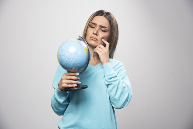 休闲穿着蓝色运动衫的金发女孩手里拿着一个地球仪 仔细地思考着 努力地记住目的地地球地球