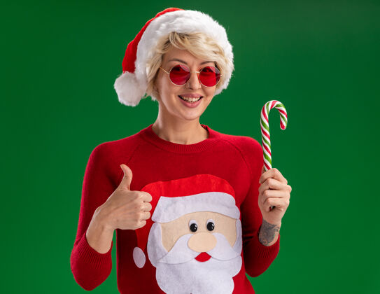 拇指快乐的年轻金发女人戴着圣诞帽和圣诞老人的圣诞毛衣戴着眼镜看着拿着圣诞糖果手杖在绿色的墙上孤立地竖起大拇指女人年轻毛衣