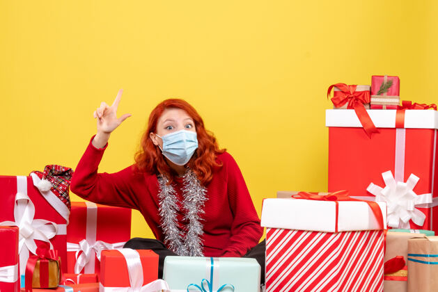 病毒黄色墙壁上戴着面具的年轻女子围坐在圣诞礼物旁的正视图礼物快乐周围