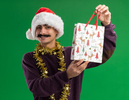 年轻快乐的小胡子男人戴着圣诞老人帽子 脖子上戴着金属丝 手里拿着圣诞礼物的纸袋 看着相机 微笑着自信地站在绿色的背景上圣诞快乐周围自信
