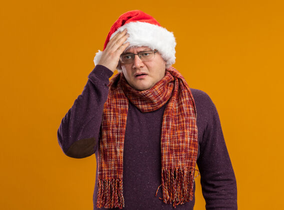周围遗憾的是 戴着眼镜 戴着圣诞帽 脖子上围着围巾 手放在头上 看着橙色背景上孤立的一面手成人眼镜