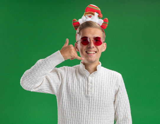 快乐快乐的年轻帅哥戴着圣诞老人的头带 戴着眼镜 在绿色的墙上做着孤立的呼唤手势圣诞老人年轻做