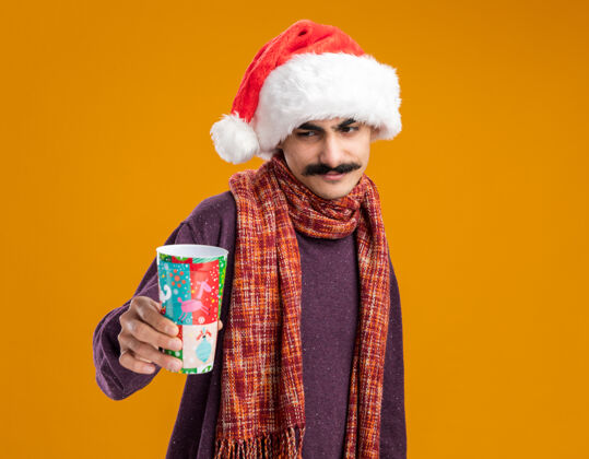 脖子快乐的胡子男人戴着圣诞老人的帽子 脖子上围着暖和的围巾 手里拿着五颜六色的纸杯 站在橙色的背景上 面带微笑地看着它圣诞节站着围巾