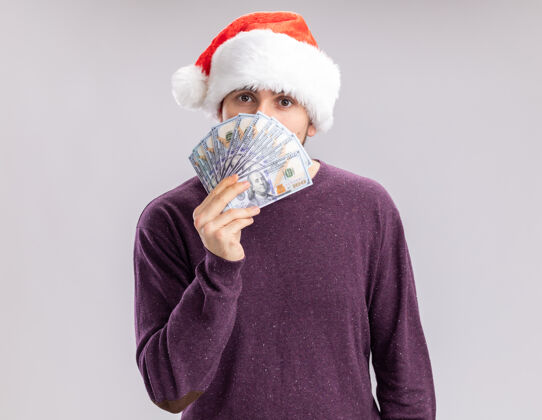 年轻人一个穿着紫色毛衣 戴着圣诞帽的年轻人 手里拿着现金 脸上盖着钱 严肃地看着相机 站在白色的背景上现金掩护圣诞老人