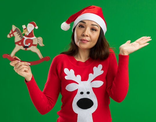 帽子戴着圣诞老人帽 穿着红色毛衣 拿着圣诞玩具的年轻女子站在绿色背景下 面带微笑地看着相机拿着毛衣微笑