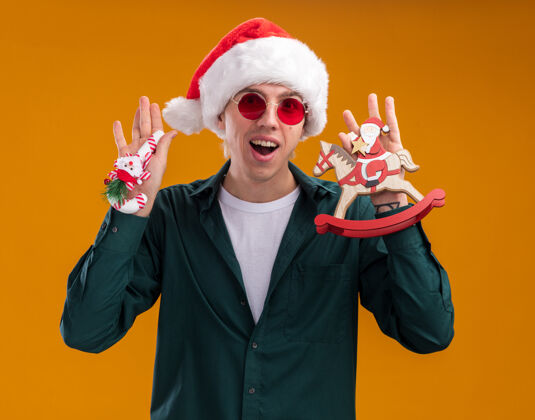 年轻令人印象深刻的年轻金发男子戴着圣诞老人帽子和眼镜举行的摇马雕像和甜蜜的藤条装饰圣诞老人看着相机孤立的橙色背景帽子装饰品摇摆