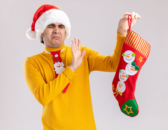 圣诞老人一个穿着黄色高领毛衣 戴着圣诞帽 打着滑稽领带的年轻人 手里拿着圣诞长袜 站在白色背景上 带着厌恶的表情看着它恶心帽子年轻