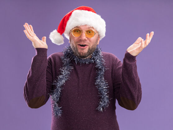 中年令人印象深刻的中年男子戴着圣诞帽 脖子上戴着金箔花环 戴着眼镜 两只空手孤立在紫色的墙上周围展示空的