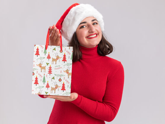 帽子穿着红色毛衣 戴着圣诞帽 手里拿着彩色纸袋 手里拿着圣诞礼物的快乐小女孩站在白色的背景下 面带微笑地看着相机毛衣微笑年轻人