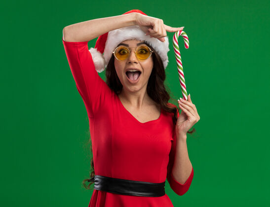 垂直令人印象深刻的年轻漂亮女孩戴着圣诞帽和眼镜手持圣诞糖果手杖垂直看着相机隔离在绿色的背景圣诞节印象深刻举行