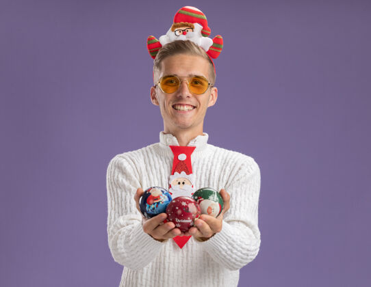 头带快乐的年轻帅哥戴着圣诞老人的头带和领带戴着眼镜拿着圣诞球饰品孤立在紫色的墙上有复制空间戴着领带快乐