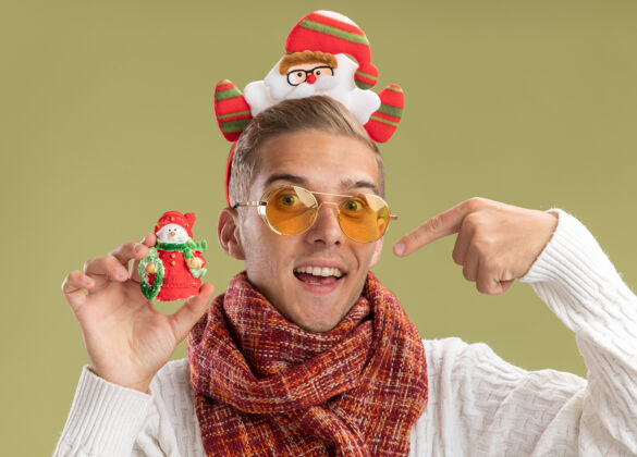 雪人给人印象深刻的年轻帅哥戴着圣诞老人的头带和围巾拿着和指着橄榄绿墙上孤立的雪人圣诞饰品戴着帅气橄榄