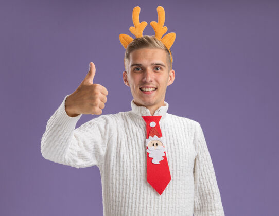 拇指快乐的年轻帅哥戴着驯鹿鹿角头带 打着圣诞老人的领带 在紫色的墙上孤立地竖起大拇指头带圣诞老人年轻