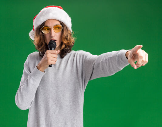 眼镜戴着圣诞老人帽 戴着黄色眼镜的年轻人对着麦克风说话 食指指着站在绿色背景上的一边圣诞老人年轻人帽子