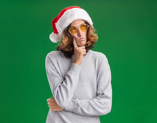 一旁戴着圣诞老人帽 戴着黄色眼镜的年轻人站在绿色的背景下 茫然地看着一边圣诞老人眼镜帽子