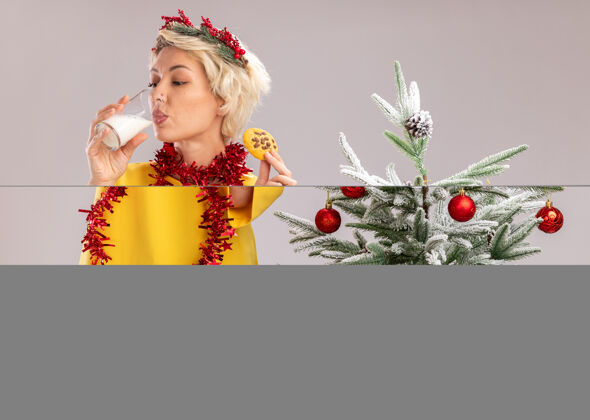 杯子年轻的金发女人戴着圣诞花环 脖子上戴着金属丝花环 站在装饰好的圣诞树旁 手里拿着一杯牛奶和饼干 喝着白色背景上隔离的牛奶喝酒饼干圣诞节