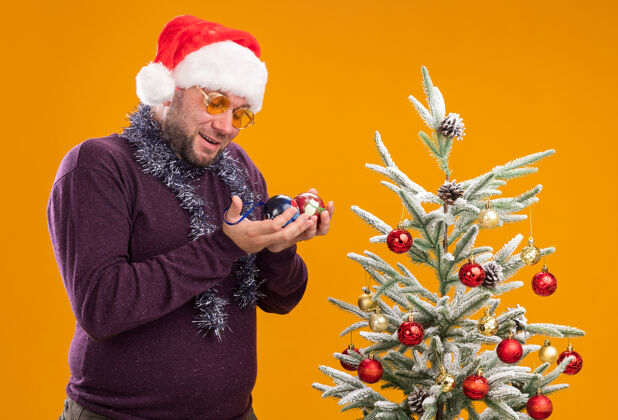 新年令人印象深刻的中年男子戴着圣诞帽 脖子上戴着金箔花环 戴着眼镜站在侧视图中 旁边抱着圣诞树印象深刻站着圣诞老人