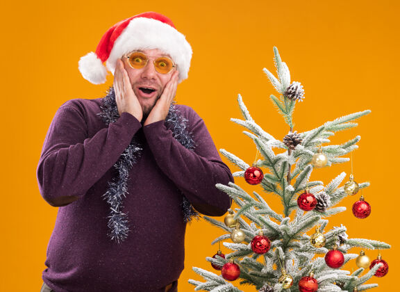 保持兴奋的中年男子戴着圣诞帽 脖子上戴着金箔花环 戴着眼镜 站在装饰好的圣诞树旁 手放在脸上 看着橙色背景上的相机帽子周围眼镜