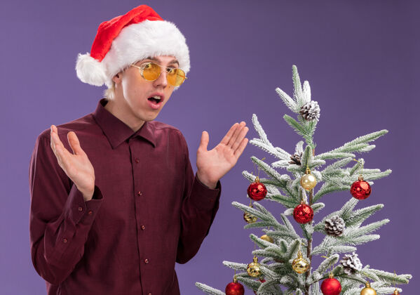 年轻令人印象深刻的年轻金发男子戴着圣诞帽和眼镜站在装饰圣诞树附近看着相机显示空手孤立在紫色的背景眼镜金发装饰