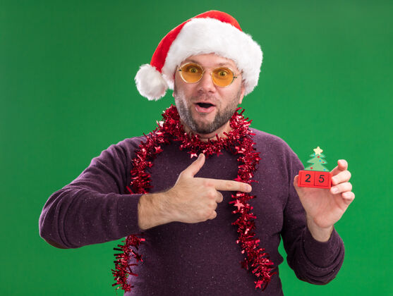 帽子令人印象深刻的中年男子戴着圣诞帽 脖子上戴着金箔花环 戴着眼镜 拿着并指着圣诞树玩具和日期 看着隔离在绿色背景上的相机日期圣诞老人脖子