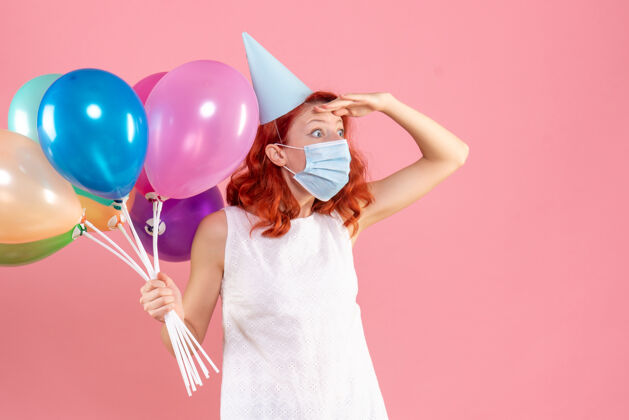 年轻前视图中的年轻女子拿着五颜六色的气球在无菌面具上粉红色的墙壁举行五颜六色聚会