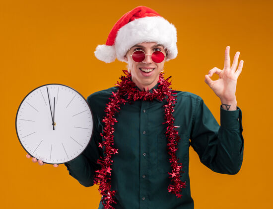 好的快乐的年轻金发男子戴着圣诞帽 戴着眼镜 脖子上戴着金属丝花环 拿着时钟看着相机 在橙色背景上做着“ok”的手势拿着脖子金发