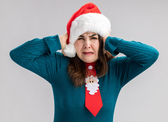 耳朵恼怒的成年白人妇女戴着圣诞帽 戴着圣诞领带 用手遮住耳朵 在白色背景上隔离 留有复印空间盖子大人帽子