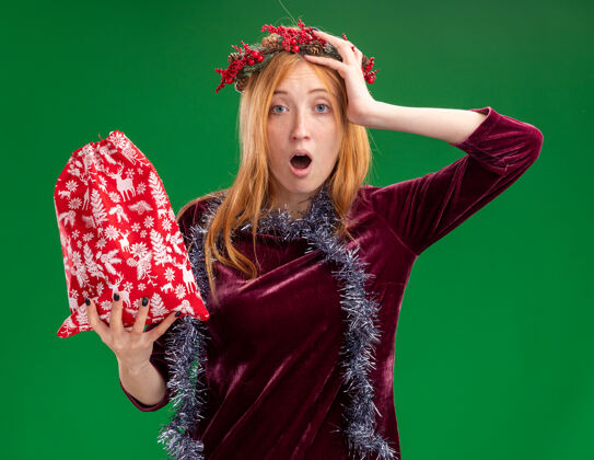 圣诞节年轻漂亮的女孩穿着红色的衣服 脖子上戴着花环和花环 手里拿着圣诞包 手放在头上 在绿色的背景下与世隔绝抱年轻女孩