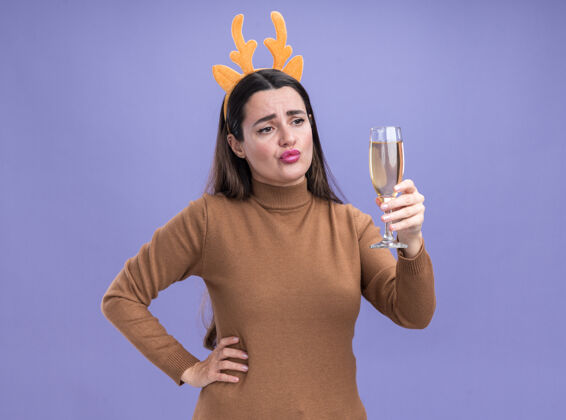 毛衣悲伤的年轻漂亮的女孩穿着棕色毛衣 抱着圣诞节的发箍 看着一杯香槟 手放在臀部 蓝色背景上孤立着放香槟箍