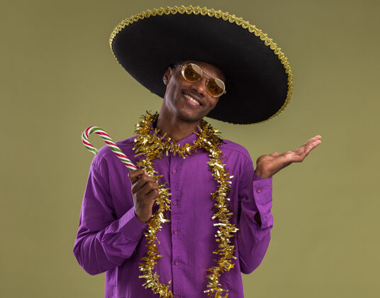 手带着墨西哥帽子和眼镜 脖子上戴着金属丝花环 手里拿着圣诞糖果手杖 面带微笑的美国黑人年轻人看着相机 在橄榄绿的背景下 一只空手孤立着金属片手杖周围