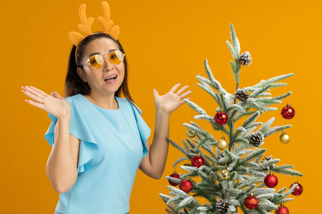 树身穿蓝色上衣的年轻女子 戴着滑稽的鹿角边和黄色眼镜 看着摄像机 站在橙色背景下的圣诞树旁 既高兴又兴奋边缘兴奋鹿
