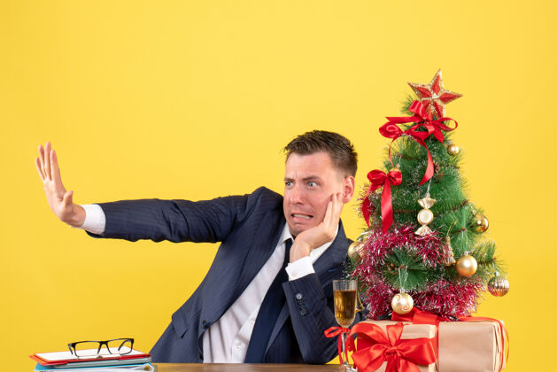 办公室前视图焦躁不安的男子试图阻止坐在圣诞树和黄色墙上的礼物附近的桌子上的东西礼物男人男人