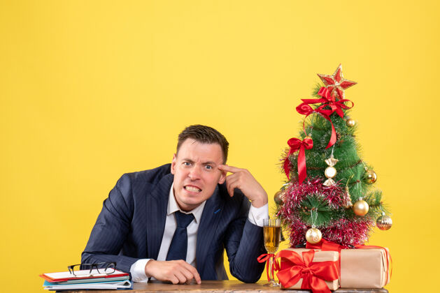 男性正面图愤怒的年轻人坐在圣诞树旁的桌子旁 黄色墙壁上免费赠送礼物西装礼物圣诞节
