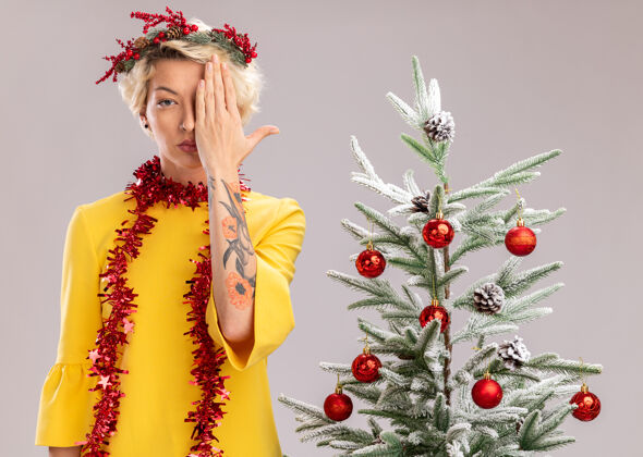 封面年轻的金发女人戴着圣诞花环 脖子上戴着金属丝花环 站在装饰好的圣诞树旁 看着相机遮住半张脸 手被隔离在白色背景上相机装饰金发