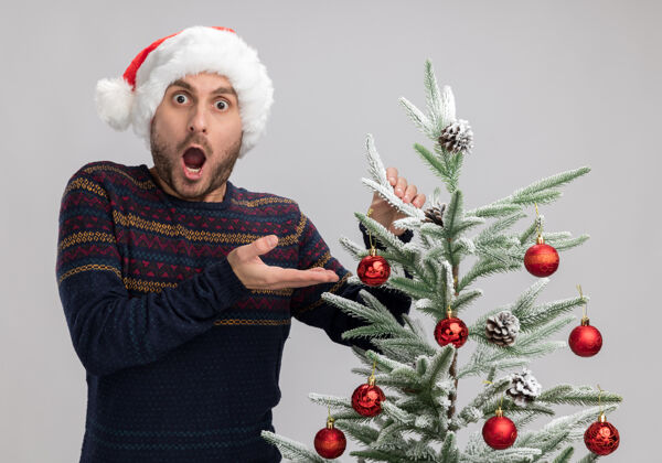 震惊震惊的年轻白种人戴着圣诞帽站在圣诞树旁抚摸着它看着隔离在白色背景下的相机相机圣诞树