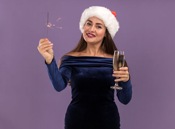 相机微笑着的年轻漂亮女孩 穿着蓝色连衣裙 戴着圣诞帽 手里拿着闪闪发光的东西 手里拿着一杯香槟 对着紫色背景上的镜头圣诞节帽子女孩