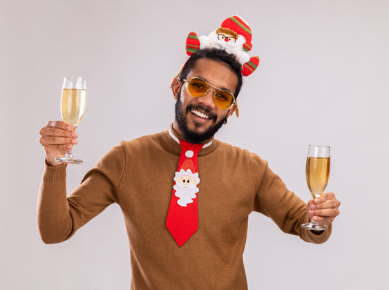 非洲非洲裔美国人 身穿棕色毛衣 头戴圣诞老人耳环 打着有趣的红色领带 手拿两杯香槟 站在白色背景上 快乐而愉快地微笑着圣诞老人搞笑微笑