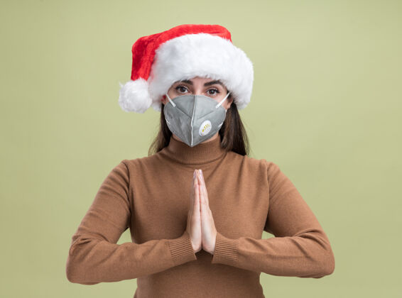 手势年轻漂亮的女孩戴着圣诞帽 戴着医用面具 在橄榄绿的背景上显示出祈祷的手势医疗帽子圣诞节