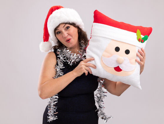 印象深刻印象深刻的中年妇女戴着圣诞帽 脖子上戴着金箔花环 抱着圣诞老人的枕头 看着隔离在白色背景上的相机圣诞老人抱抱枕头