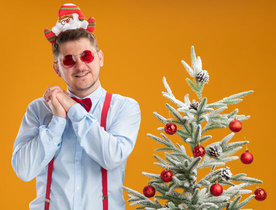 惊喜快乐的年轻人穿着吊带领结 戴着圣诞老人和红眼镜 站在圣诞树旁 手牵着手 在橙色背景下等待惊喜眼镜站立圣诞老人
