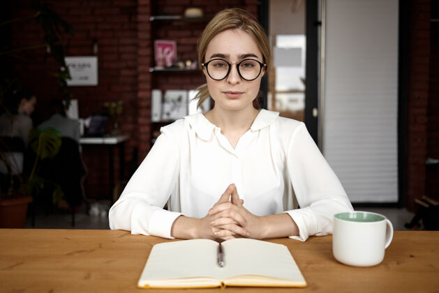 专业照片中 一位自信友好的年轻女性人力资源经理身穿白色上衣 戴着眼镜 在面试时双手紧握 坐在办公桌前 认真地提问和倾听沟通工作面试职业