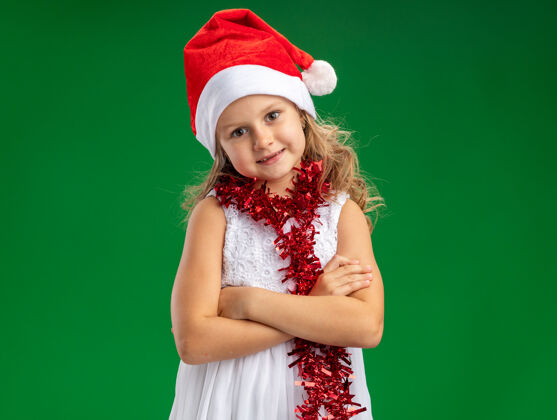 女孩高兴的歪着头的小女孩戴着圣诞帽 脖子上戴着花环 双手交叉 背景是绿色的交叉小圣诞节