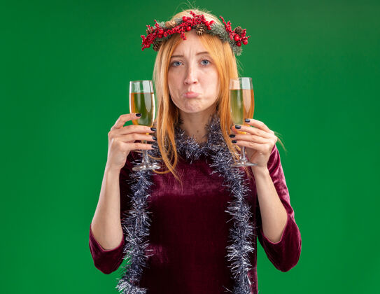 抱着悲伤的年轻漂亮的女孩穿着红色的裙子 脖子上戴着花环和花环 手里拿着一杯香槟 背景是绿色的杯子脖子香槟