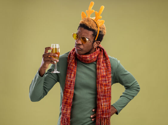 玻璃站在绿色背景上 一个非洲裔美国人 戴着滑稽的边 戴着鹿角 脖子上围着围巾 手里拿着一杯香槟 兴致勃勃地看着它举行好奇搞笑
